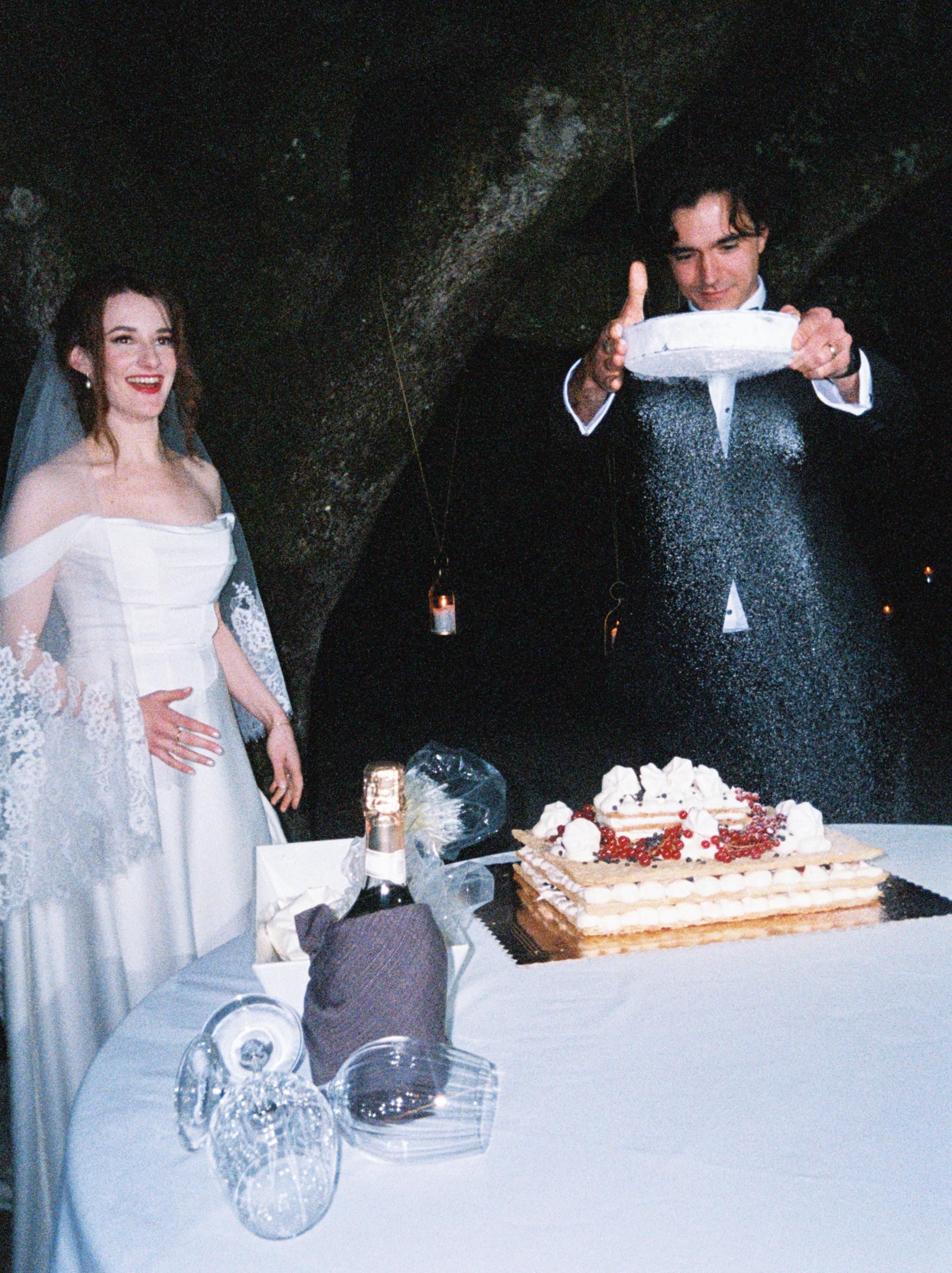A couple celebrates at their wedding reception table with a decorated cake and white decor in a dimly lit venue.