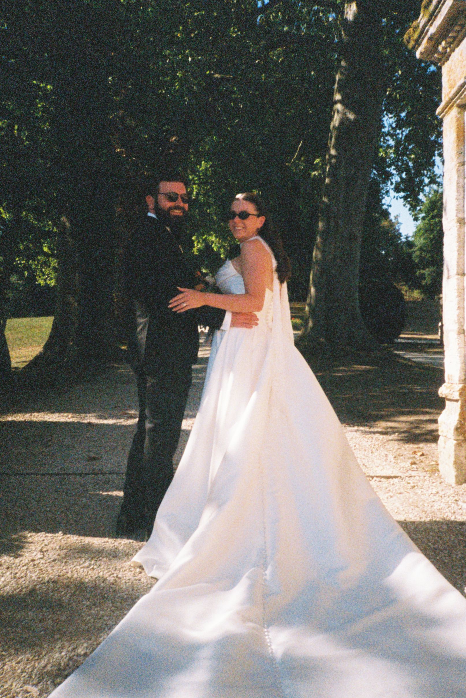 A couple in wedding attire dances and spins together in a sunlit garden setting with trees in the background.