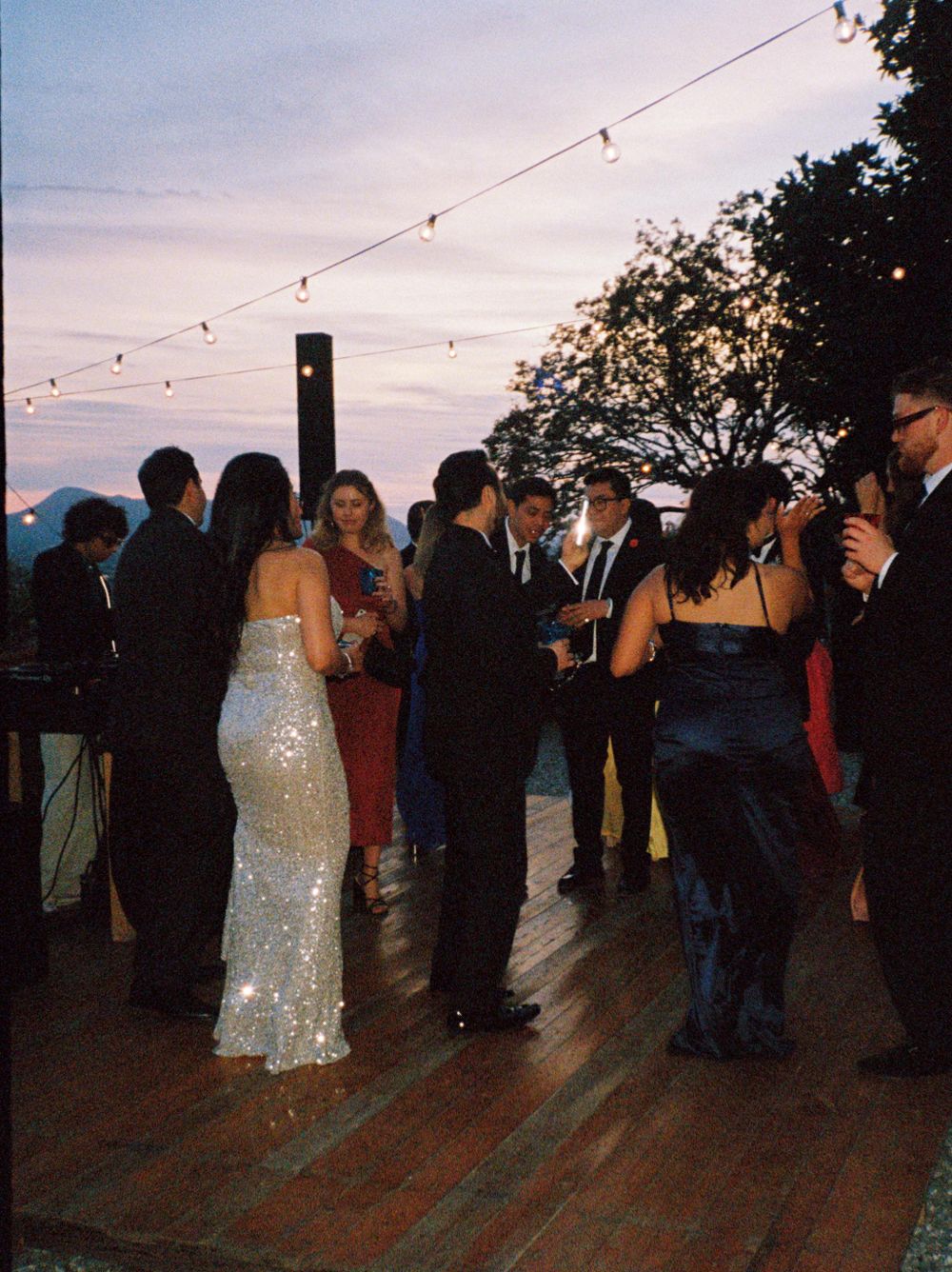 Evening wedding reception with guests dancing on outdoor deck under string lights.