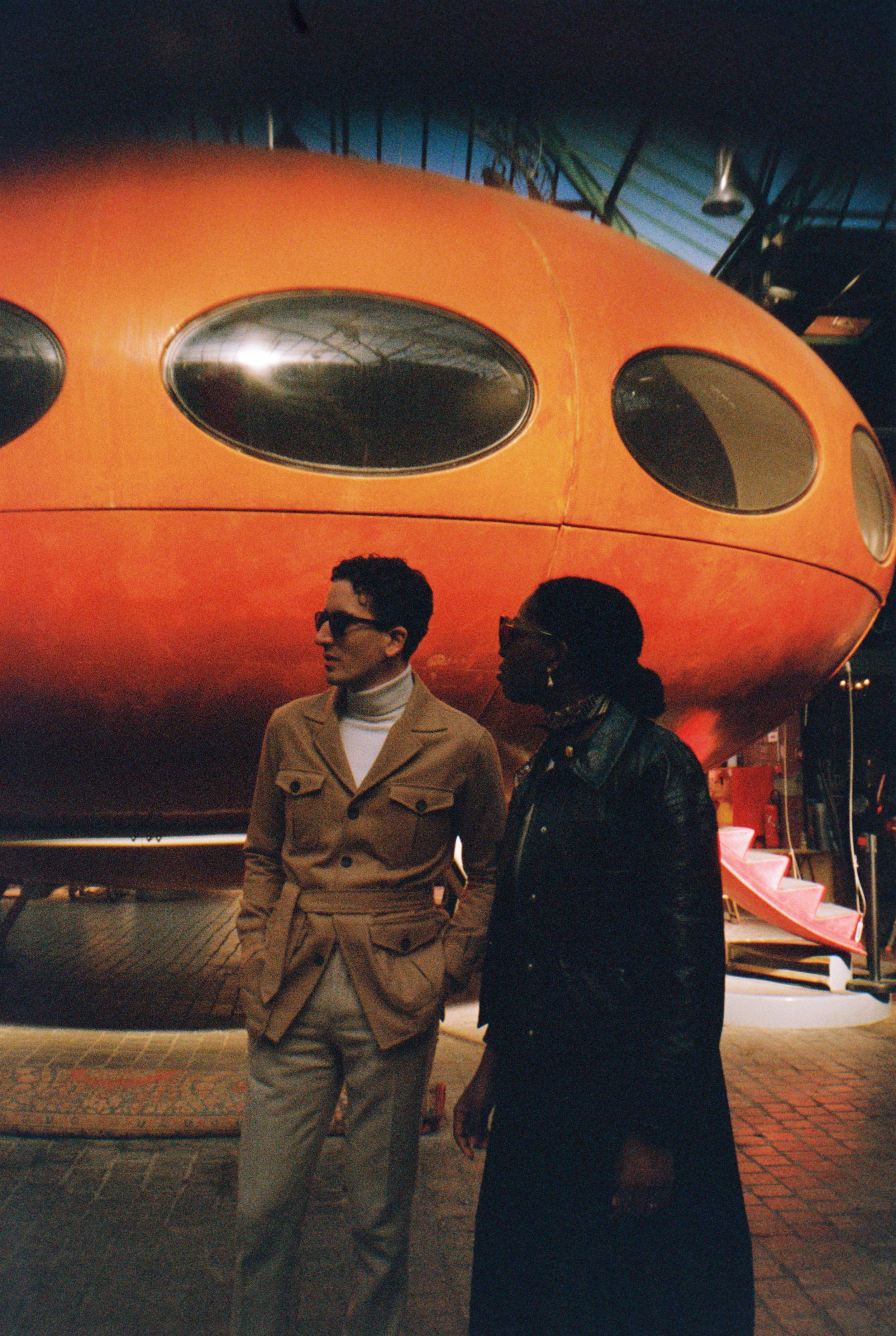 Two people standing next to a retro orange Futuro house with circular windows.