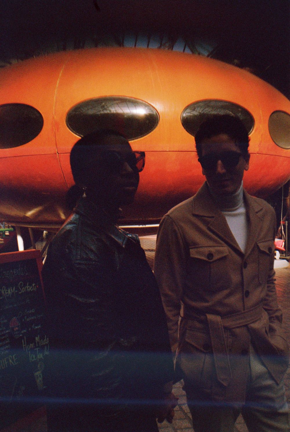 Two figures in silhouette against an orange retro-futuristic building at dusk.