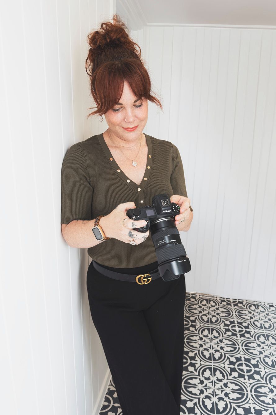 Photographer in olive green top and black pants holds professional camera while standing on patterned tile floor.