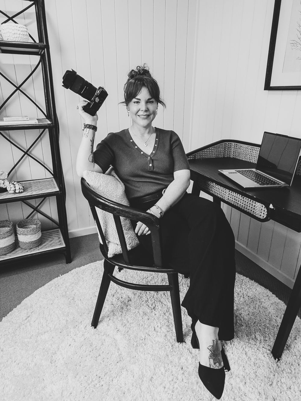 Black and white photo of a photographer sitting in a vintage chair with a camera, in a home studio setting.
