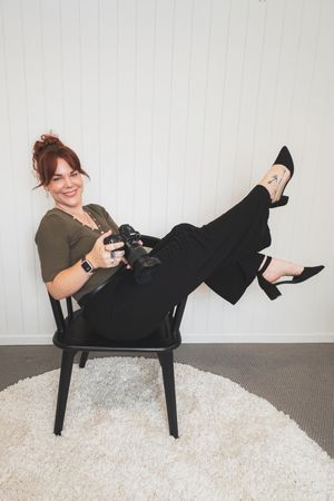 A photographer in an olive sweater and black pants sits casually in a black chair on a white rug while holding a camera.