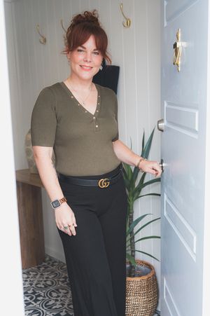 A woman in an olive green polo shirt and black pants poses against a white wall with a patterned floor.