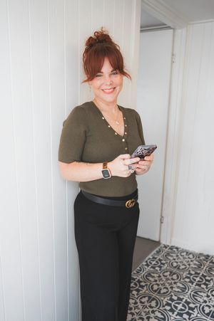 Woman in olive green top and black skirt stands smiling while checking her phone in a room with patterned flooring.