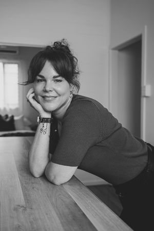Black and white portrait of someone leaning on a wooden table with a warm smile in casual attire.