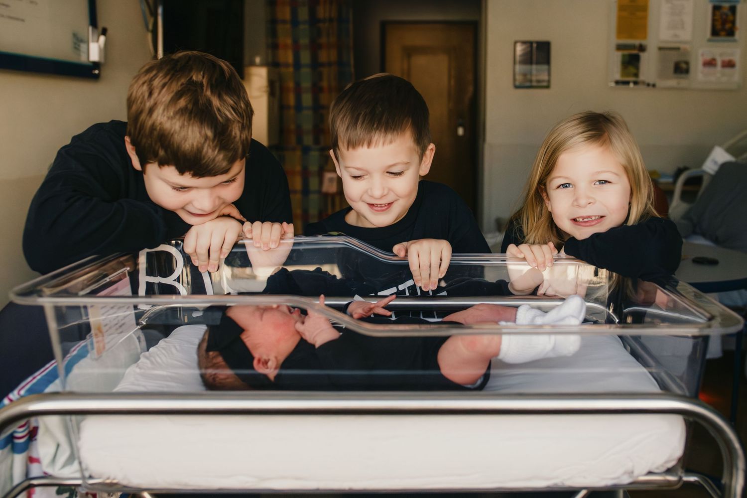 Siblings looking at their newborn sister in hospital bassinet. Captured by Maryland Photographer Mallory Bramlett.