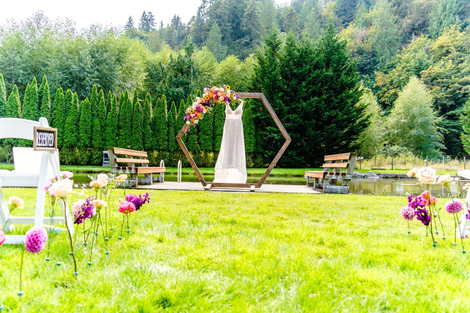 Outdoor wedding ceremony setup with hexagonal arch, floral decorations, and wooden benches surrounded by evergreen trees.