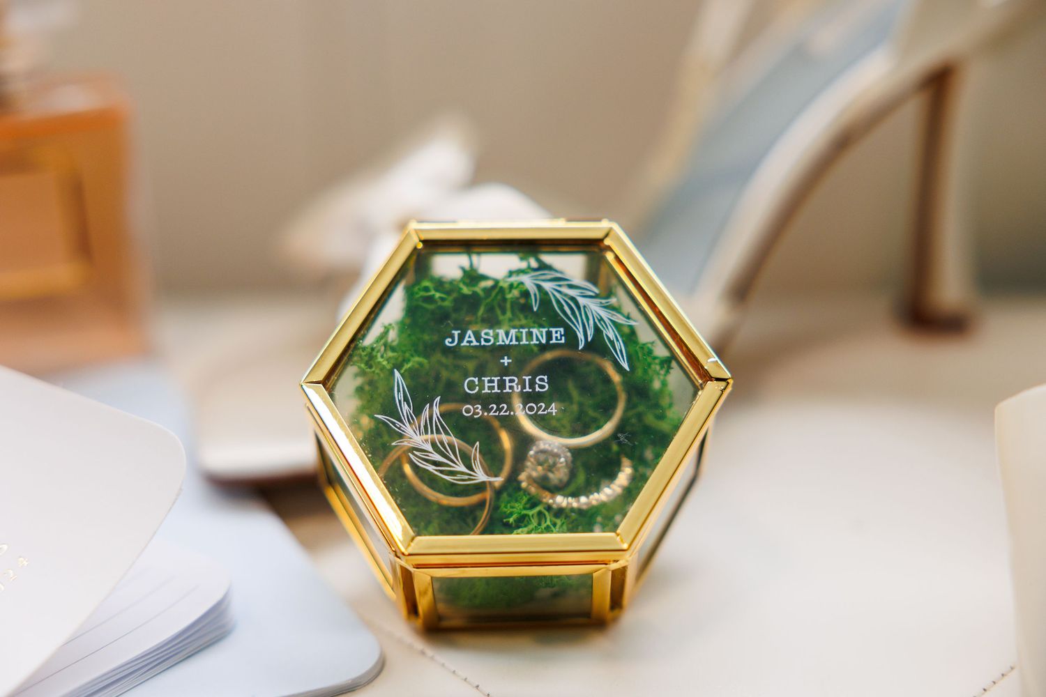 A small hexagonal glass ring box with gold trim contains wedding rings nestled in green velvet.