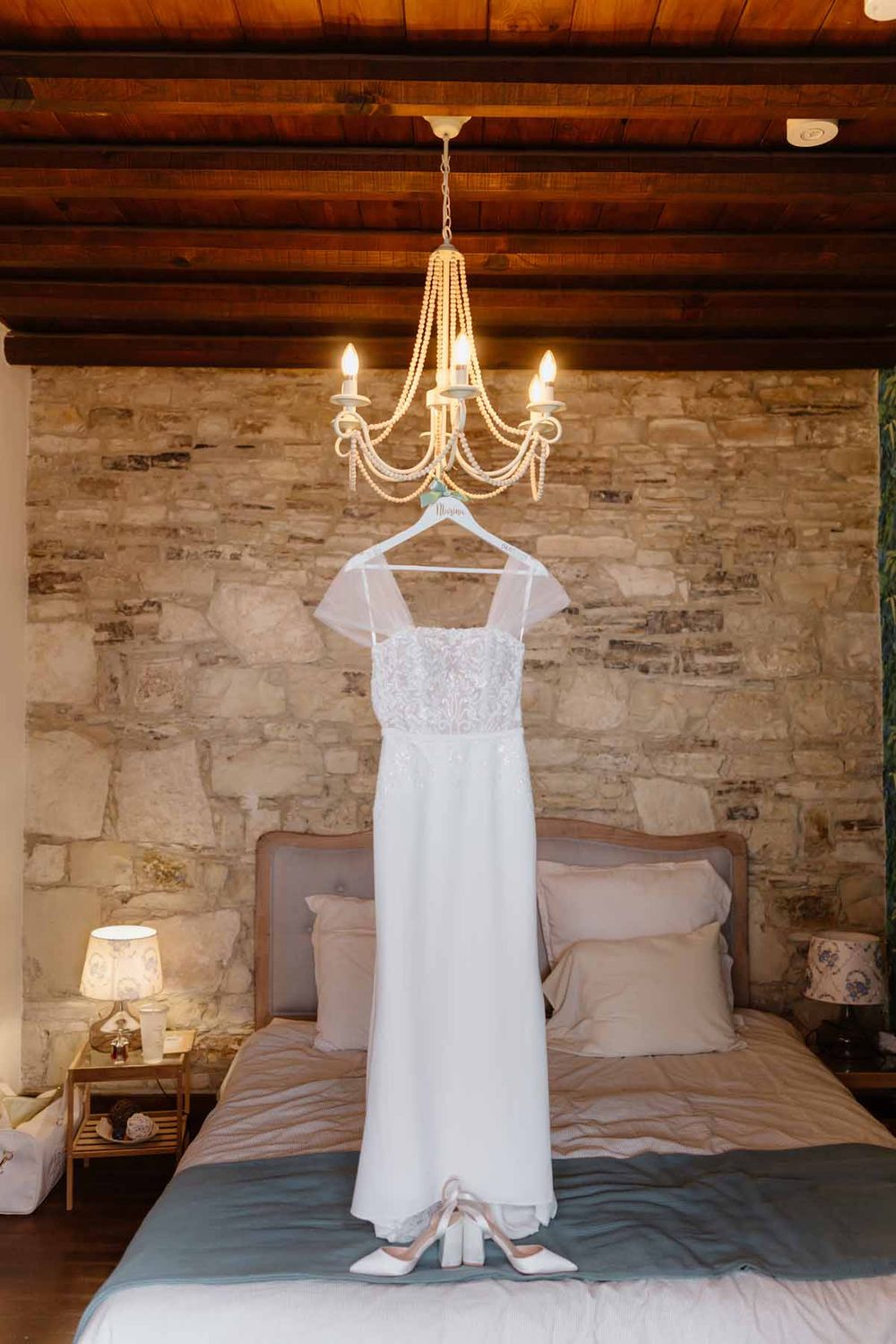 White wedding dress displayed on a hanger beneath an elegant chandelier in a rustic bedroom with stone walls and wooden beams.
