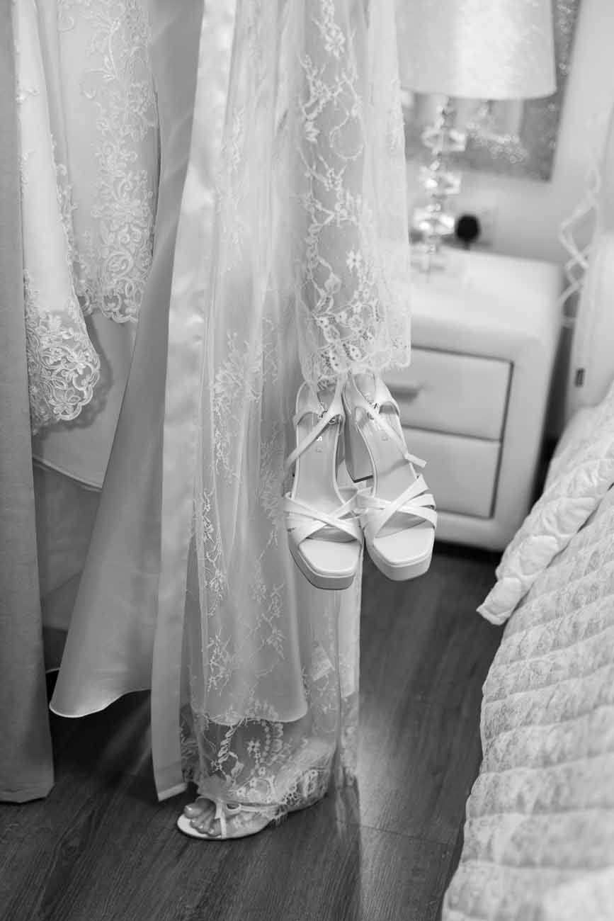 Black and white photo of elegant bridal heels hanging beside lace wedding gowns in a bridal boutique.