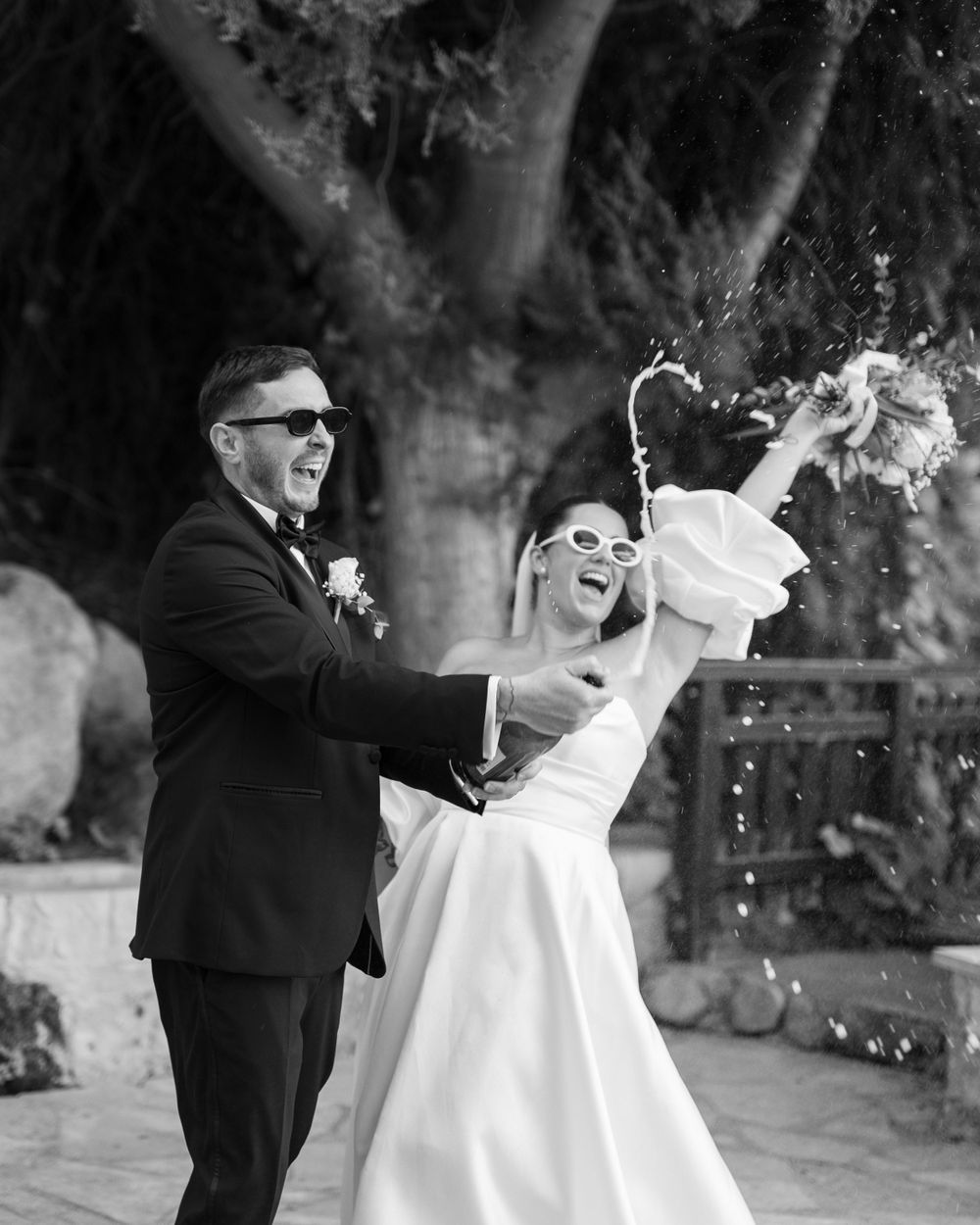 Joyful newlyweds celebrate with champagne spray during outdoor wedding photoshoot in black and white.