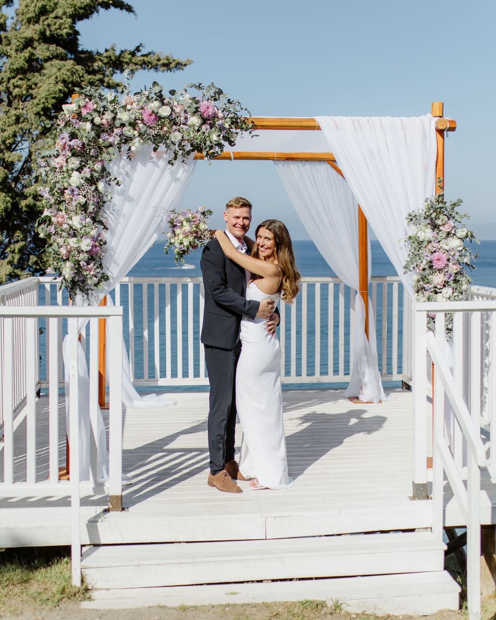 A romantic wedding ceremony takes place under a white draped arch decorated with pink and purple flowers overlooking the ocean.