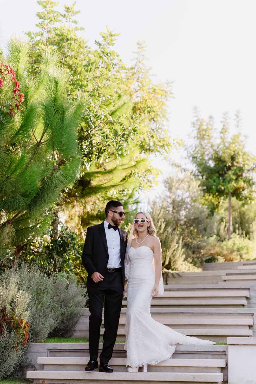 Bride and groom pose on outdoor steps surrounded by lush greenery wearing sunglasses on wedding day.