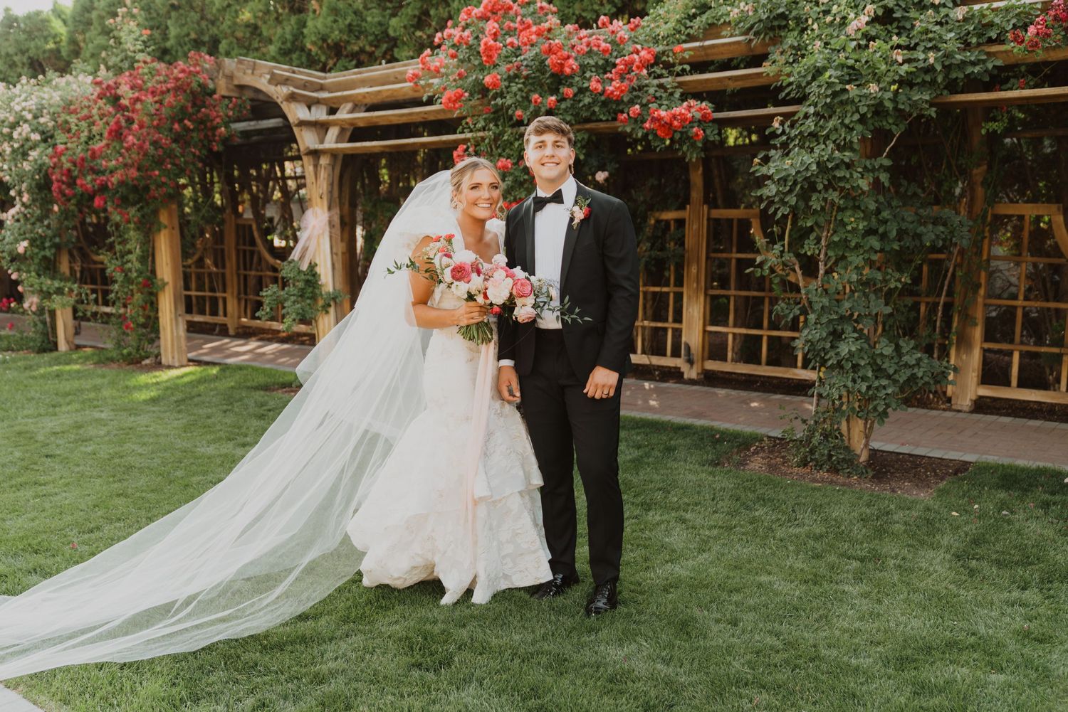 Wedding portrait in front of wooden pergola with red climbing flowers.