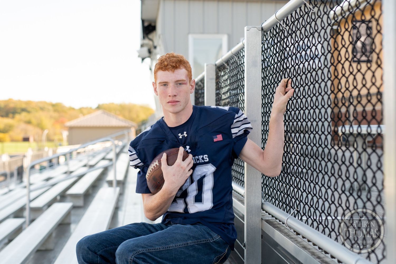 Football player in jersey leans against chain link fence at stadium.