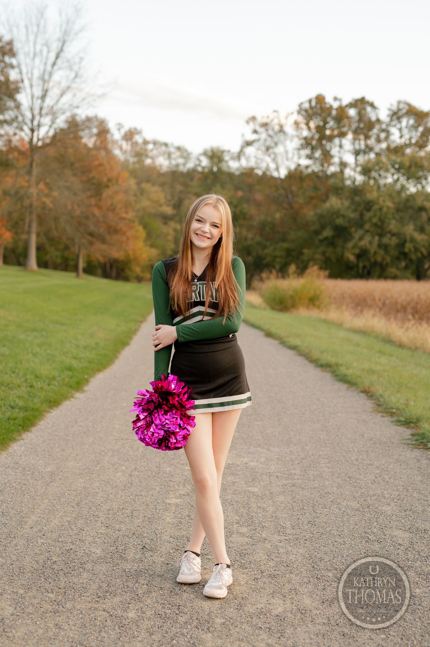 Cheerleader smiles while holding pink pom poms on a pathway with fall foliage in the background.
