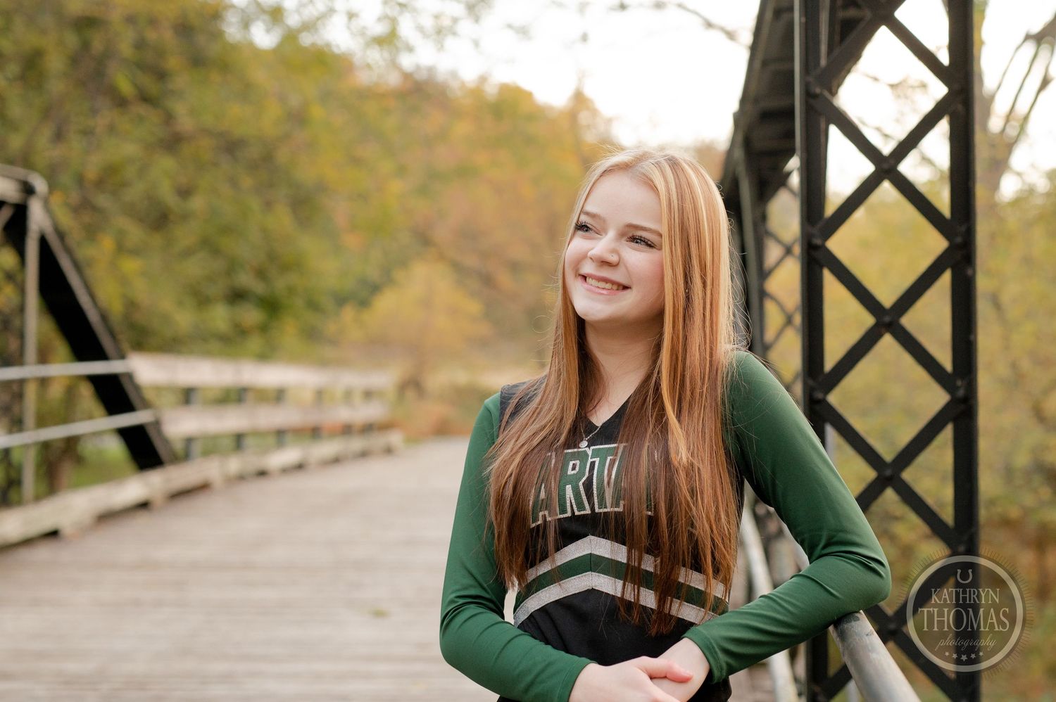 Cheerleader in green and black uniform poses on rustic bridge with autumn scenery behind.