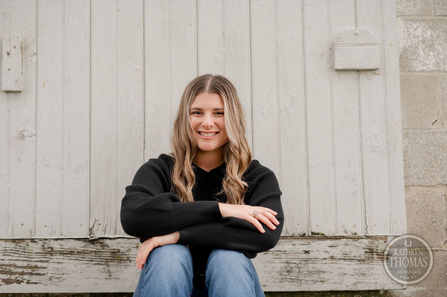 Relaxed portrait against white barn wall wearing black sweater and blue jeans.
