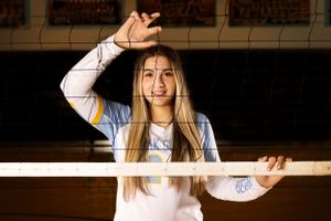 Volleyball player posing behind net wearing white jersey while reaching up to block in gymnasium.