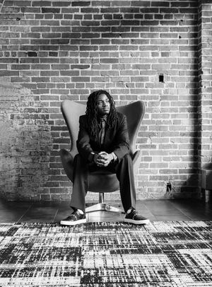 Person with dreadlocks sitting in a stylish chair against a brick wall, black and white portrait photography.