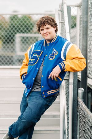 A smiling teen in a blue and gold varsity jacket with a music note patch leans on bleacher railings outdoors.