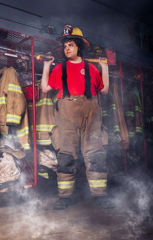 Firefighter in turnout pants and red shirt poses with axe on shoulders in smoky fire station gear room.