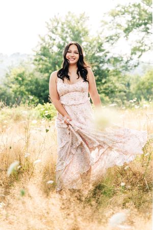 Flowing floral dress catches the breeze in a sunlit meadow with golden grass and green trees in background.