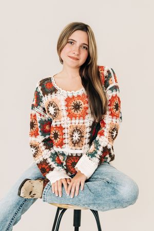 Young woman in colorful crochet granny square sweater and jeans sits on stool against neutral background.