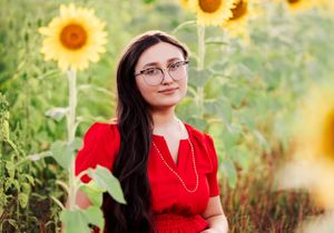 Young woman in red dress with glasses posing among vibrant sunflowers in a lush green field.