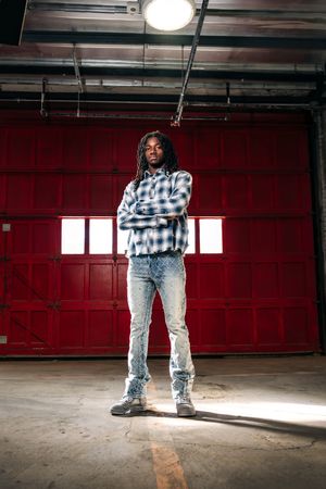 Man with dreadlocks in plaid shirt and jeans stands confidently with arms crossed in front of red garage door.