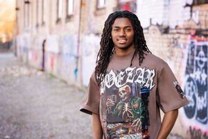 Young man with dreadlocks wearing a graphic tee poses in front of a graffiti wall outdoors.