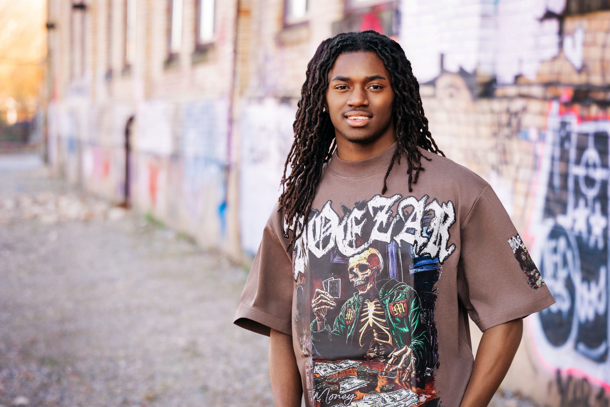 Young man with dreadlocks wearing a graphic tee poses in front of a graffiti wall outdoors.