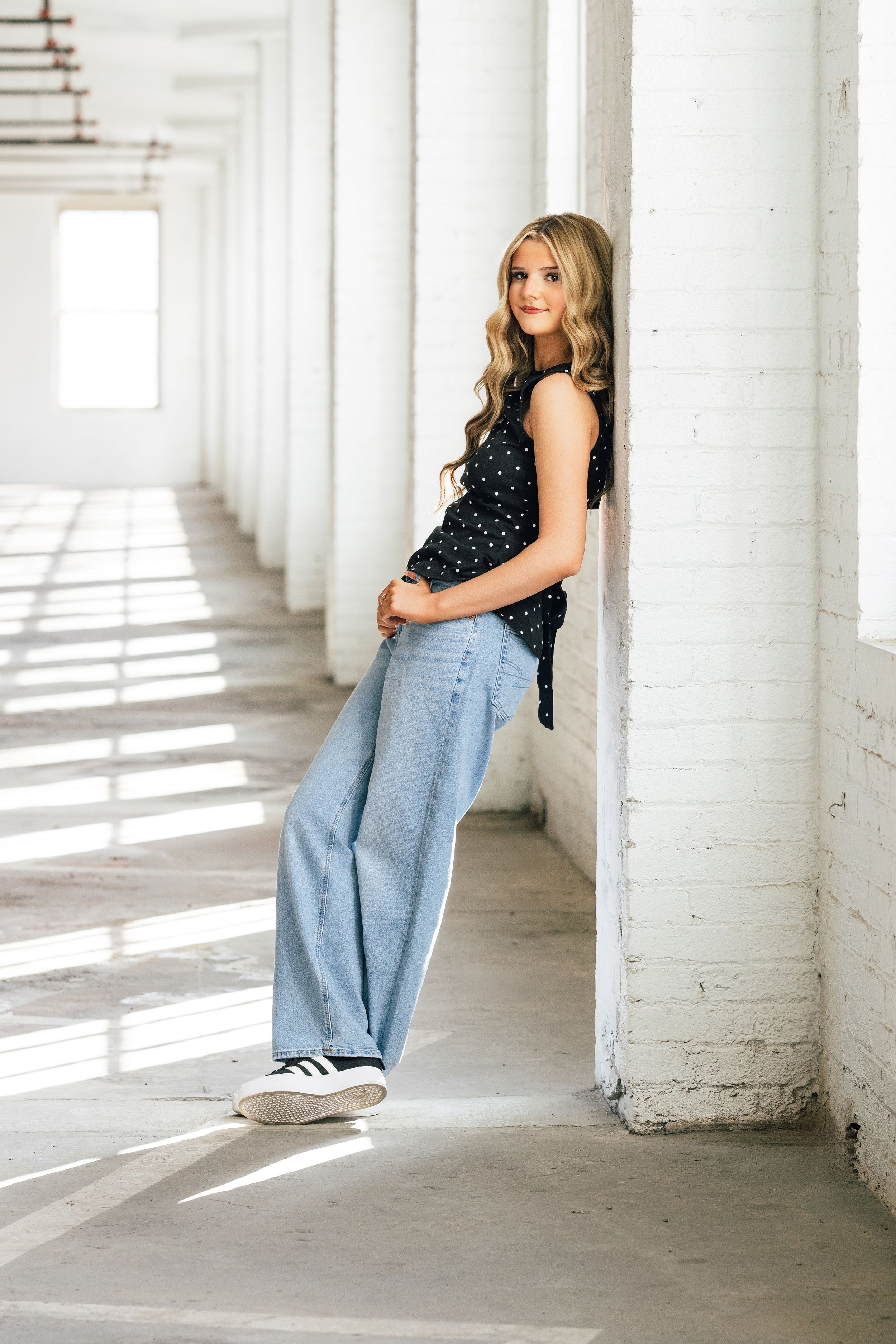 Young woman in wide-leg jeans and black polka dot top leans against white brick column in bright hallway.