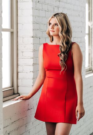 Young woman in a red sleeveless mini dress posing by a white brick wall near a window.