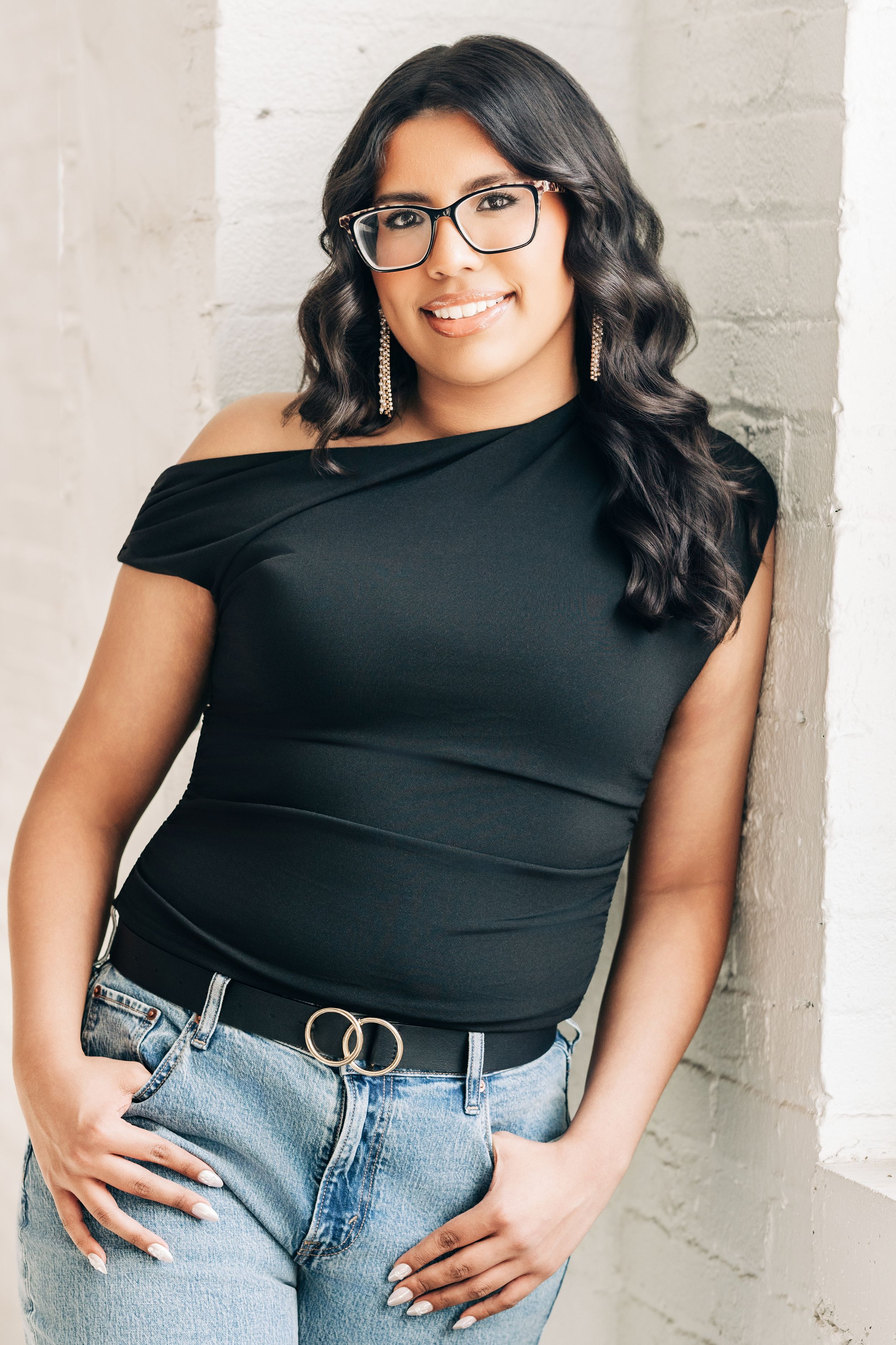 Woman in black off-shoulder top and jeans smiling against white brick wall, wearing glasses and gold earrings.