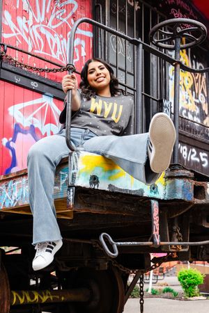 Young woman in WVU shirt and jeans sits on vintage train car surrounded by colorful graffiti walls, smiling.
