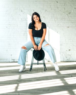 Person in ripped jeans and black top poses casually on chair against white brick wall in modern studio setting.