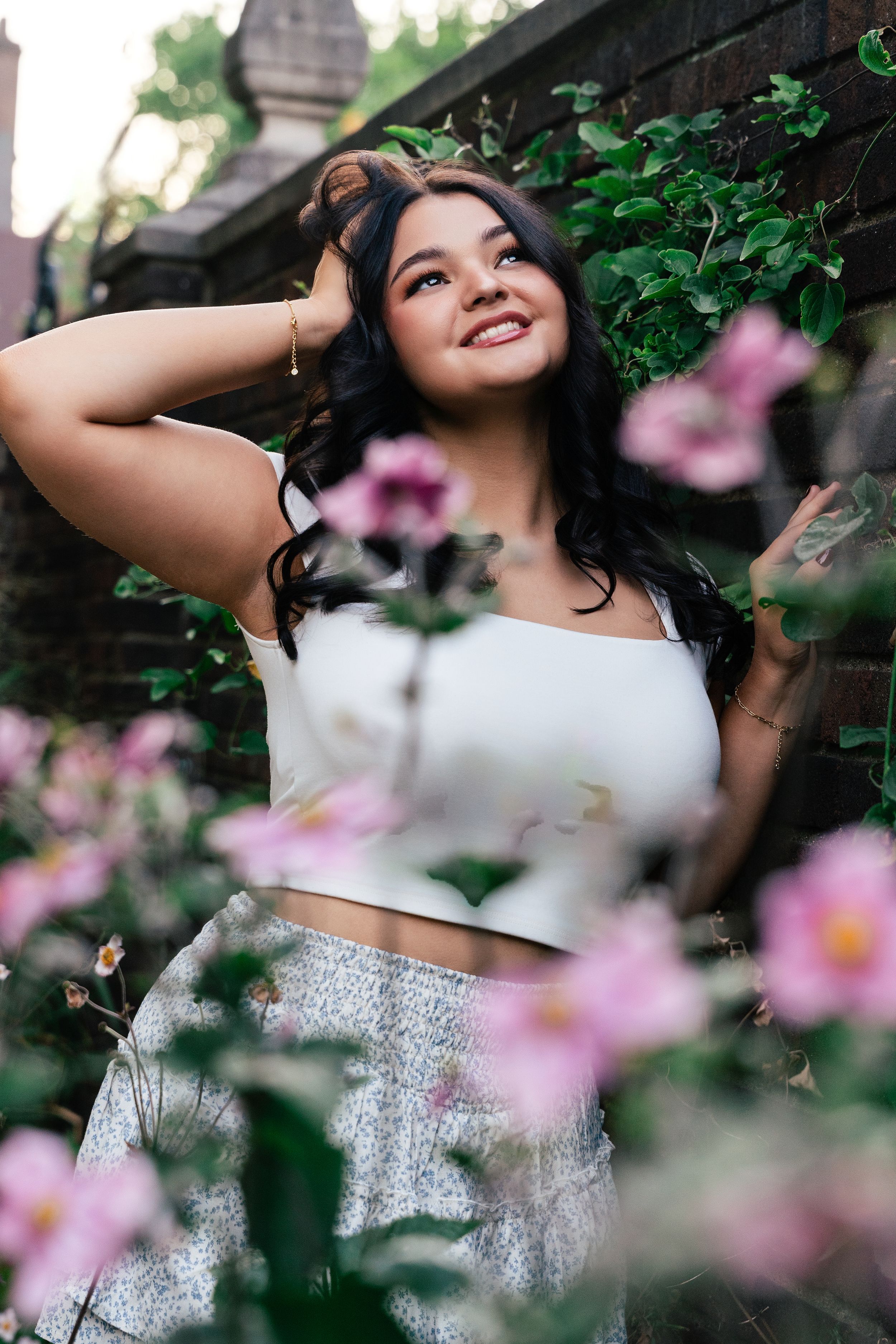 A person in a white crop top and sparkly skirt poses gracefully among pink cherry blossoms in a garden setting.
