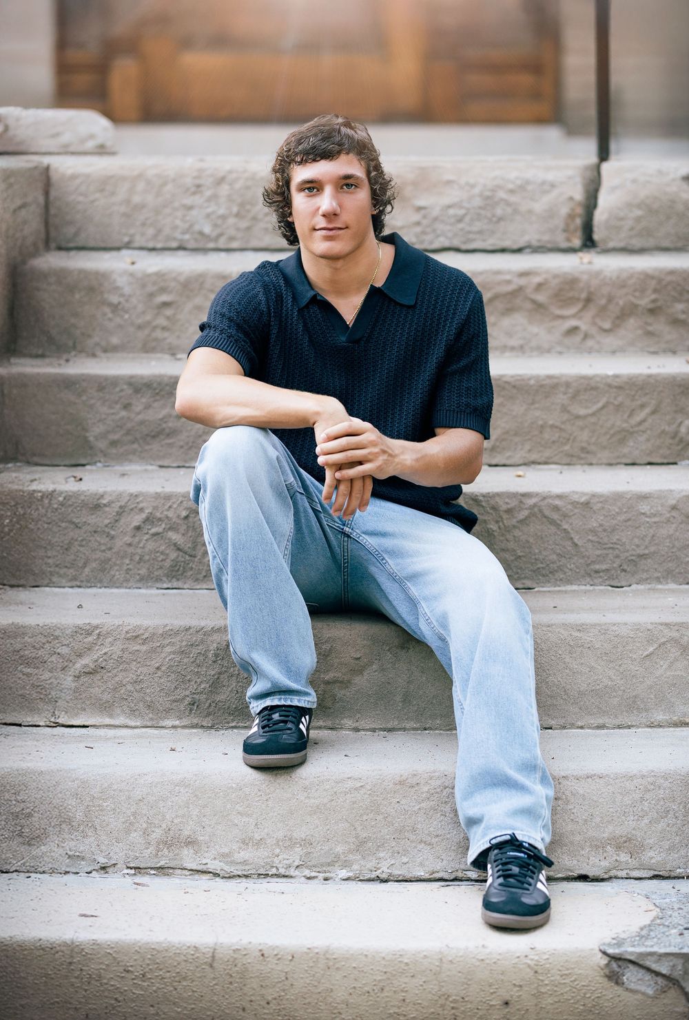 Young man with curly hair sitting on stone steps wearing a black polo shirt and light blue jeans.