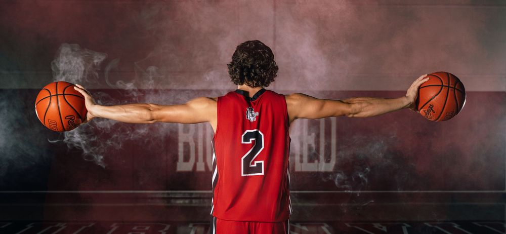 Basketball player in red jersey #2 holds two basketballs with arms outstretched against a smoky dark background.