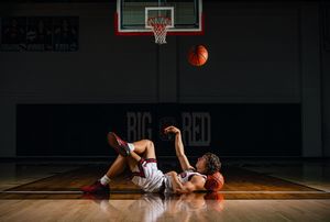 Basketball player lying on court floor with ball in air near hoop in dark gym with Big Red signage.