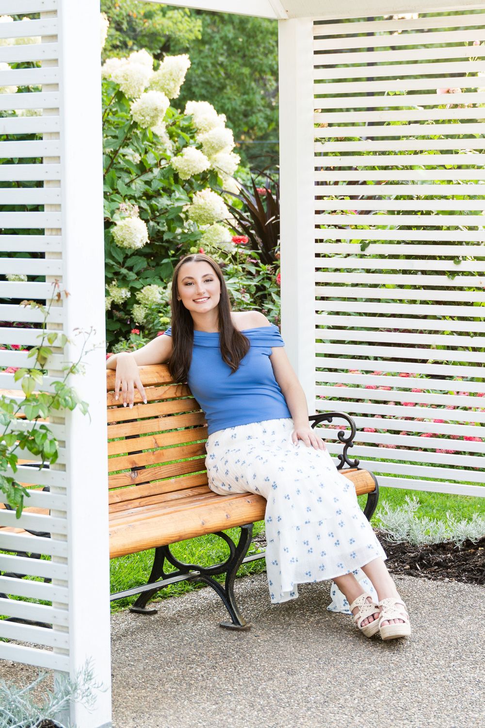 A smiling brunette woman in a blue top and floral skirt sits relaxed on a wooden bench in a lush garden.