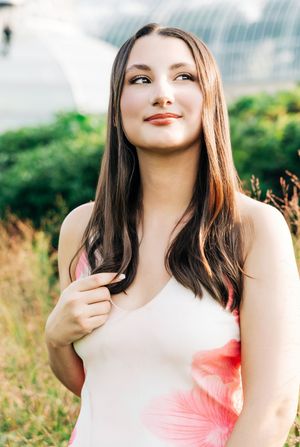 Young woman with long brown hair in a floral dress smiling outdoors with greenery in the background.