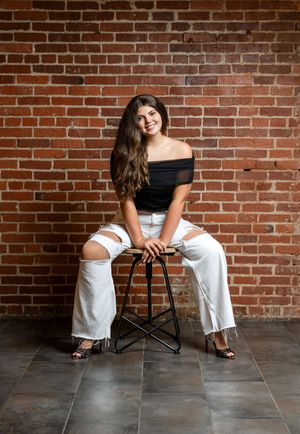 Young woman in black off-shoulder top and ripped white jeans sits on stool against rustic brick wall.
