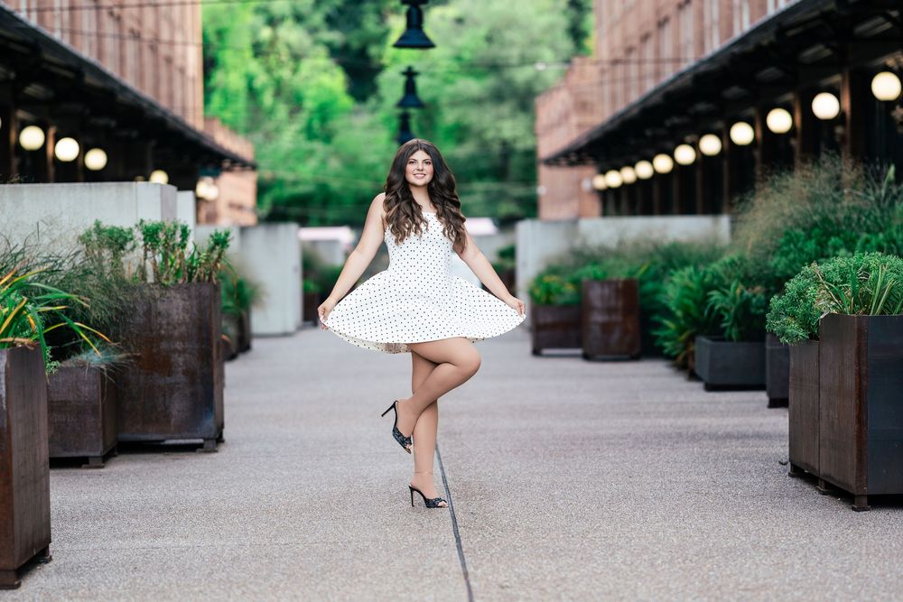Young woman in white polka dot dress twirling in elegant outdoor corridor with string lights and greenery.