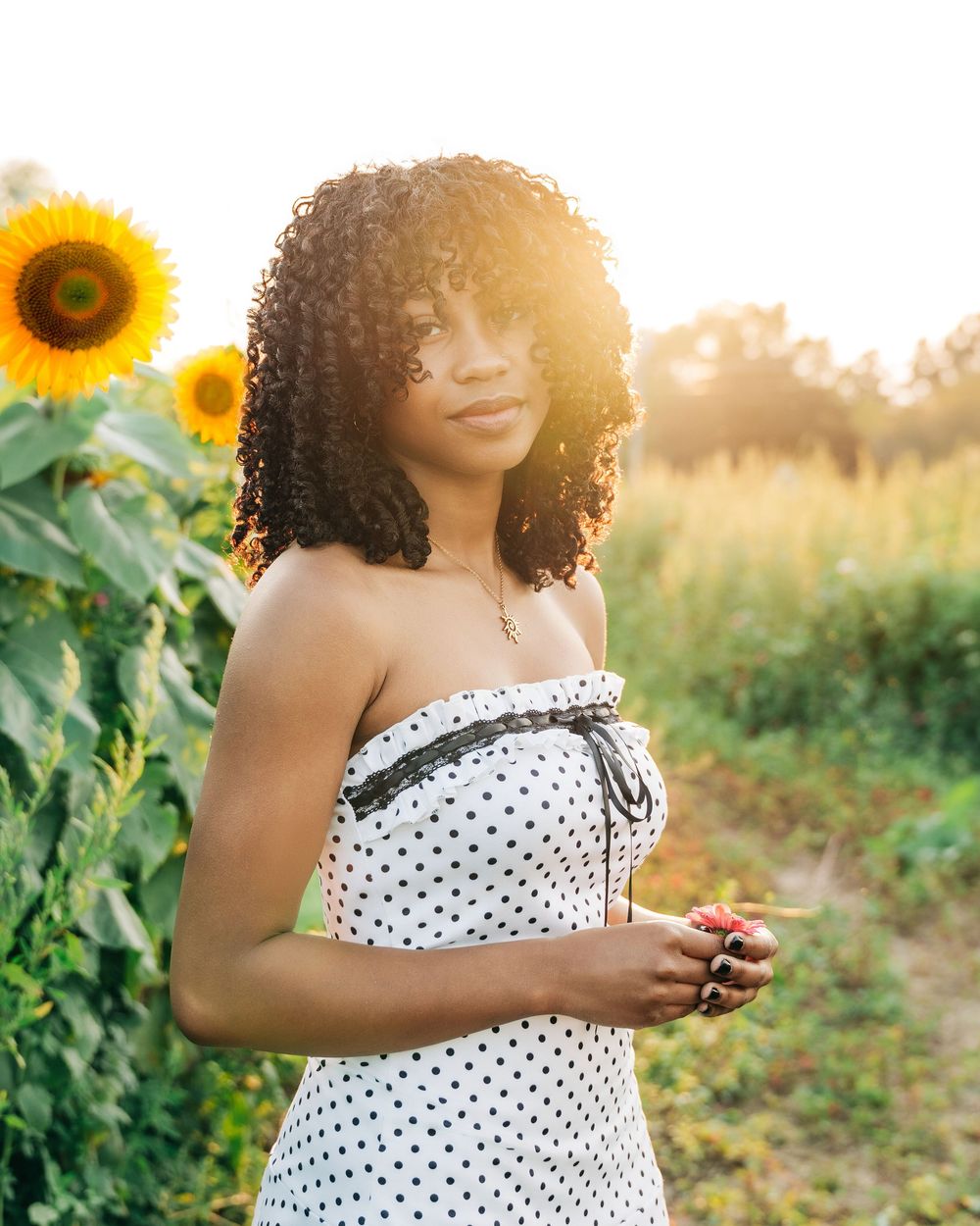 Young Black woman in polka dot strapless dress poses in sunflower field during golden hour sunset.