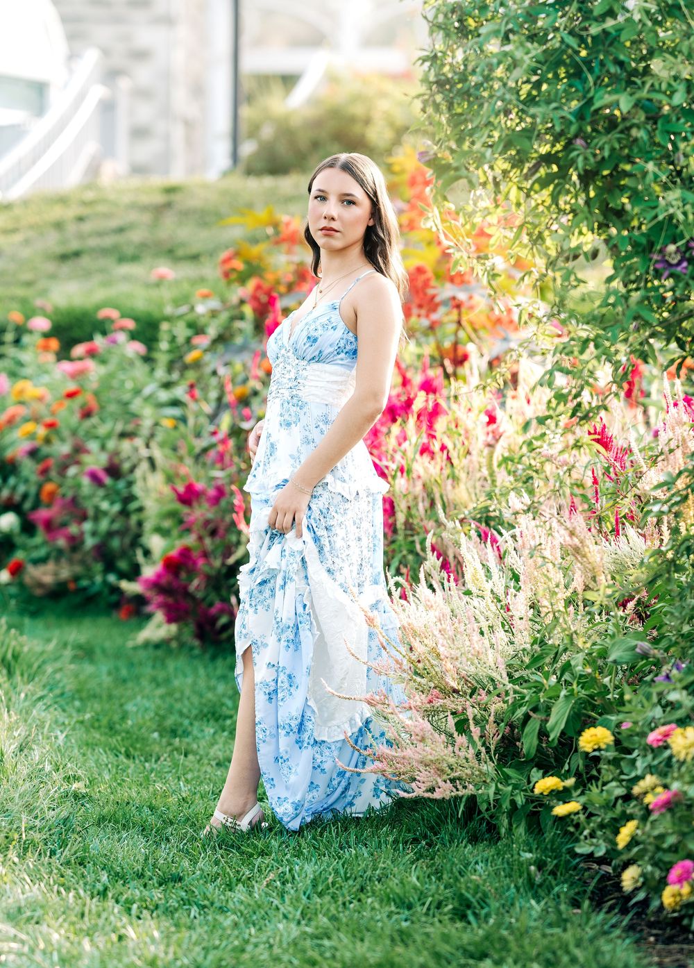 Young woman in a blue floral maxi dress posing in a vibrant garden with colorful blooms on a sunny day.