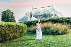 Woman in floral maxi dress standing in lush garden with glass greenhouse dome at sunset.